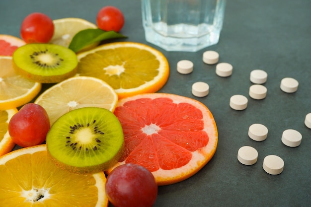 Suppléments ou aliments naturels sains Pilules éparpillées vitamines Agrumes tranchés orange citron kiwi raisins verre avec de l'eau sur un fond gris pierre
