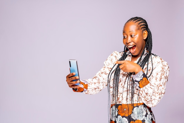 Surexcité hipster femme africaine noire pointant sur son téléphone avec sa bouche grande ouverte. Jeune femme africaine à l'aide de SMS dans les médias sociaux debout sur un fond de studio blanc