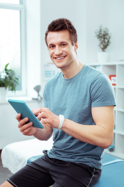 Surfer sur Internet. Jeune homme joyeux tenant sa nouvelle tablette en attendant son médecin