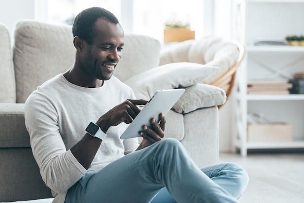 Surfer sur le net. Beau jeune homme africain touchant sa tablette numérique et souriant assis sur le tapis à la maison