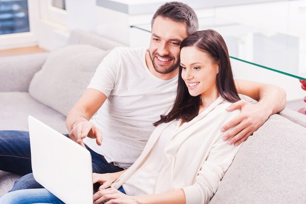 Surfer sur le net ensemble. Beau jeune couple d'amoureux se liant les uns aux autres et regardant l'ordinateur portable avec le sourire alors qu'il était assis sur le canapé ensemble