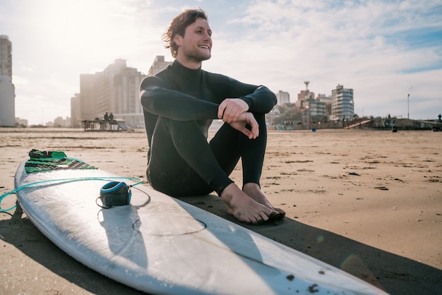 Surfeur assis sur une plage de sable et à côté de la planche de surf.