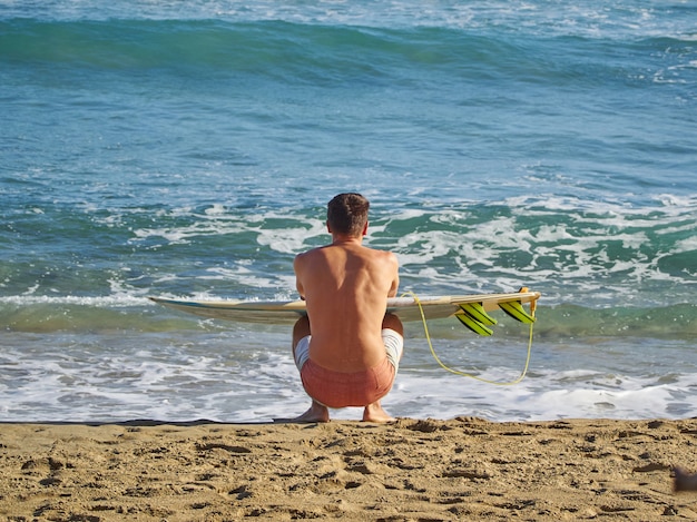 Un surfeur est assis sur la plage avec une planche de surf et regarde les vagues.