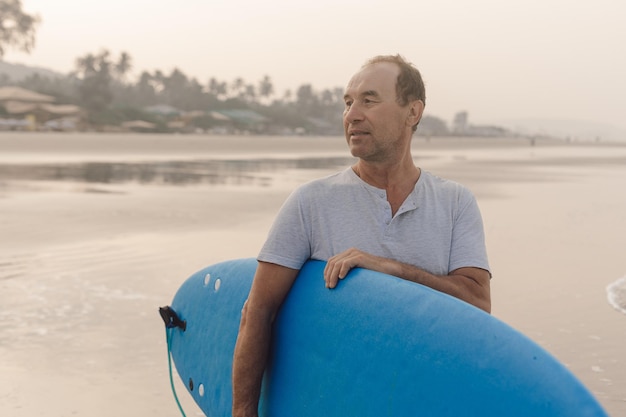 Photo surfeur mature et confiant marchant le long d'une côte vide au coucher du soleil, transportant une planche de surf bleue sous l'aisselle et regardant de côté