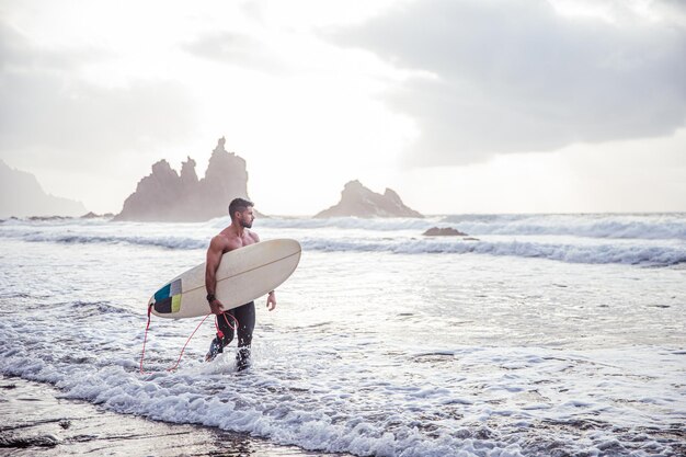 Un surfeur regarde l'océan en se promenant sur la plage avec sa planche de surf