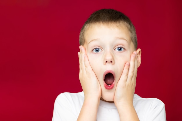 Surpris adolescent 7-10 en t-shirt blanc, avec la bouche ouverte et les mains sur les joues sur fond rouge