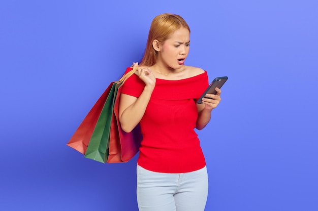 Surpris belle femme asiatique en robe rouge tenant des sacs à provisions, utilisant un téléphone portable regardant la caméra isolée sur fond violet