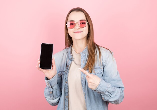 Surpris brune heureuse en veste en jean montrant l'écran du smartphone vierge et pointant avec le sourire sur le mur rose