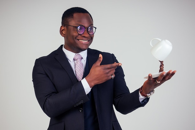 Surpris et étonné, bel homme d'affaires afro-américain dans un costume classique noir et des lunettes tenant une tasse avec des éclaboussures de grains de café volants sur une assiette sur fond blanc tourné en studio. Boisson magique du matin