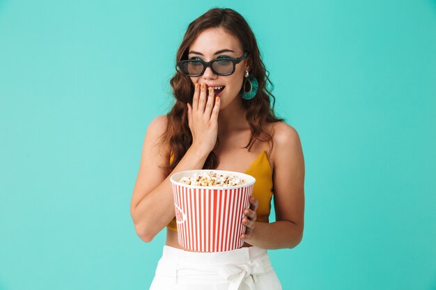 Surpris femme brune portant des lunettes tenant seau avec pop-corn et regardant la caméra