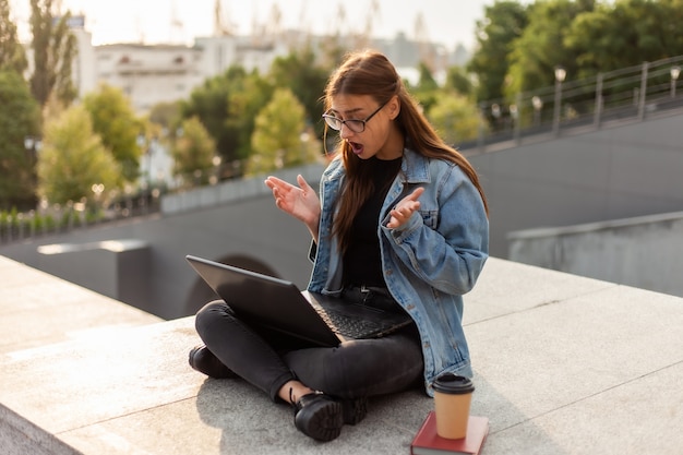 Surpris femme étudiante moderne dans une veste en jean, assis dans les escaliers, regarde l'ordinateur portable à écran Apprentissage à distance. Concept de jeunesse moderne.