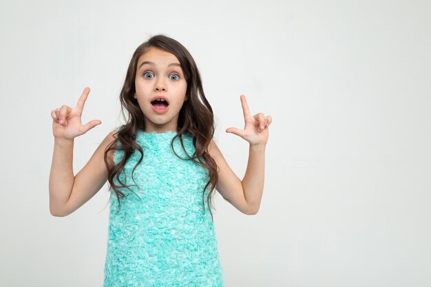 Surpris fille adolescente dans une robe bleue pouces vers le haut sur une publicité sur un fond de studio blanc.