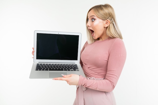 Surpris fille blonde en vêtements roses est titulaire d'un ordinateur portable avec l'affichage vers l'avant avec un vide vide sur un fond de studio blanc
