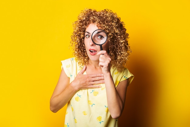 Surpris fille bouclée regarde à travers une loupe sur une surface jaune