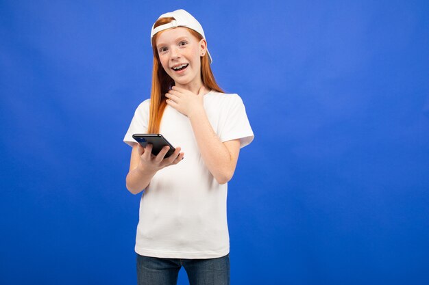 Surpris fille rousse adolescente dans un T-shirt blanc avec un gadget pour la communication en mains bleu