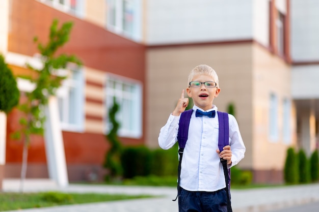 Surpris garçon blond écolier dans des verres avec un sac à dos se tient à l'école et montre un coup de pouce