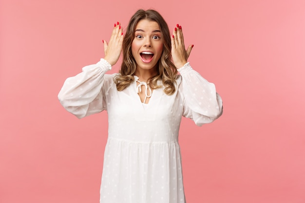 Surpris heureuse, excitée, une fille du jour b en robe blanche ouvre les yeux pour voir un incroyable cadeau incroyable d'amis à la fête, debout sur un mur rose, étonné et joyeux