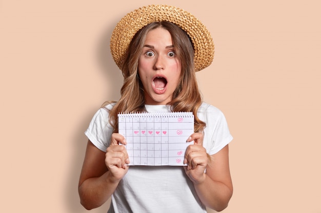 Surpris jeune belle femme à la peau pure, ouvre largement la bouche, vêtue d'un chapeau de paille et d'un t-shirt, détient un calendrier d'époque, exprime le choc, isolée sur le mur du studio beige. Concept Omg