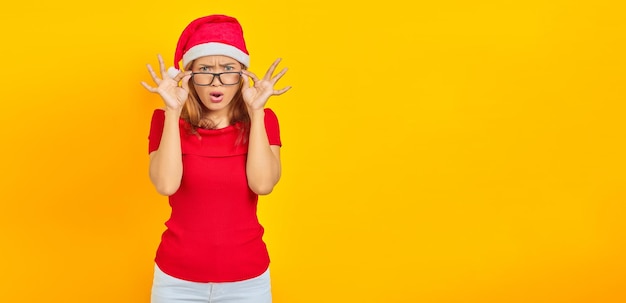 Surpris jeune femme asiatique portant un chapeau de Noël avec des lunettes tandis que la bouche ouverte sur fond jaune