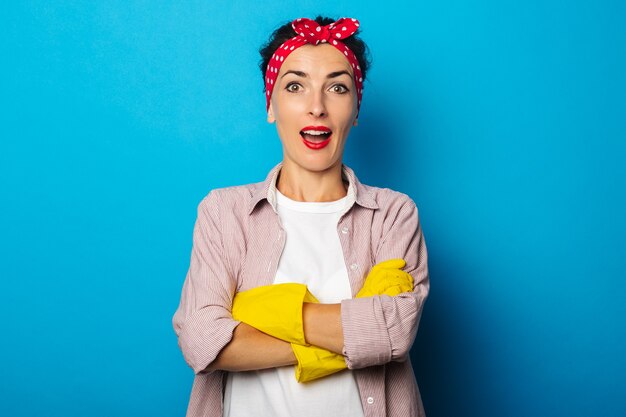 Surpris jeune femme avec un bandage sur ses cheveux croisant ses mains dans des gants de nettoyage sur une surface bleue