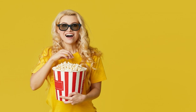 Surpris de jeune femme blonde à lunettes 3d et chemise jaune manger du pop-corn, regarde un film choquant au cinéma. Isolé