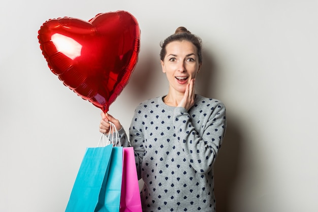Surpris jeune femme dans un pull détient des paquets pour faire du shopping et un ballon à air coeur sur un fond clair. Concept de la Saint-Valentin. Bannière.
