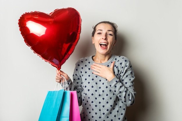 Surpris jeune femme dans un pull détient des paquets pour faire du shopping et un ballon à air coeur sur un fond clair. Concept de la Saint-Valentin. Bannière.