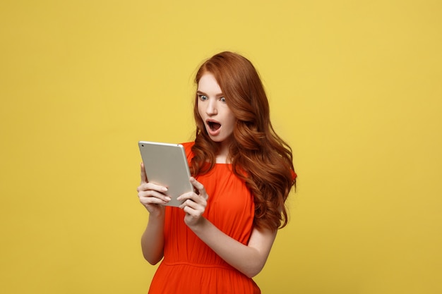 Surpris de jeune femme portant des vêtements de robe orange à l&#39;aide de tablet pc isolé sur fond jaune.