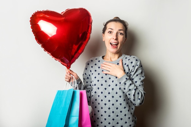 Surpris, une jeune femme en pull tient des colis pour faire du shopping et une montgolfière cardiaque sur fond clair. Concept de la Saint-Valentin. Bannière.