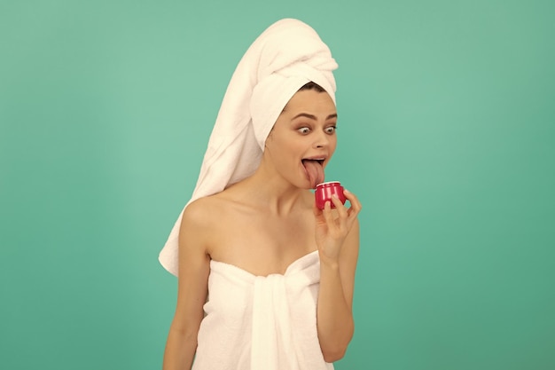 Surpris jeune femme en serviette léchant la crème pour la peau sur fond bleu