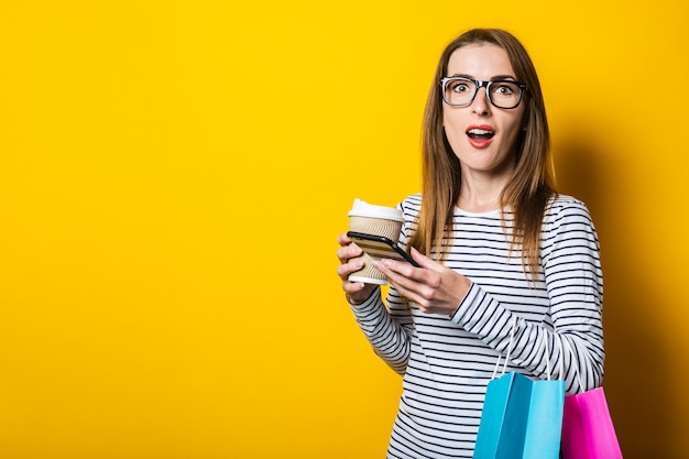 Surpris jeune femme avec un téléphone, avec des sacs à provisions, avec un sur fond jaune