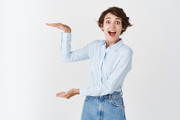 Surpris jeune femme en tenue professionnelle montrant un objet de grande taille et un regard étonné impressionné par la quantité façonnant une grande boîte debout sur fond blanc