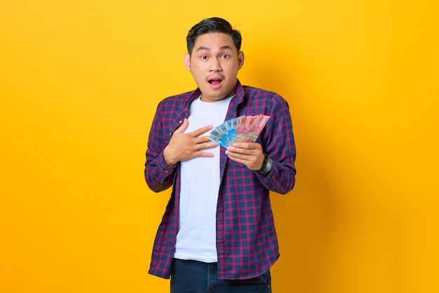Surpris jeune homme asiatique en chemise à carreaux tenant des billets de banque avec la bouche ouverte isolé sur fond jaune