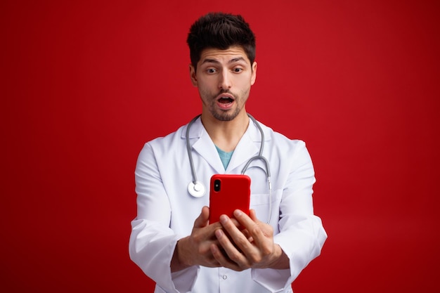 Surpris jeune médecin de sexe masculin portant un uniforme médical et un stéthoscope autour du cou tenant un téléphone portable parlant par appel vidéo isolé sur fond rouge