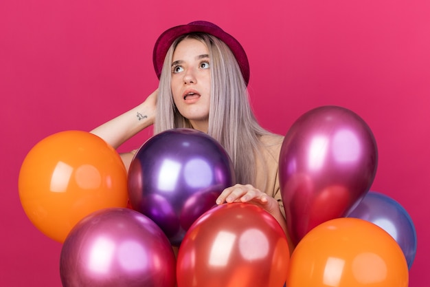 Surpris en levant la belle jeune fille portant un chapeau de fête avec des appareils dentaires debout derrière des ballons isolés sur un mur rose