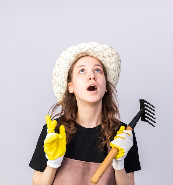 Surpris en levant le jeune jardinier portant un chapeau de jardinage avec des gants tenant un râteau isolé sur un mur blanc