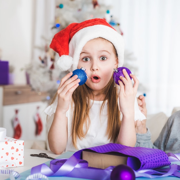 Surpris petite fille en bonnet de Noel tenant des boules bleues et violettes à Noël