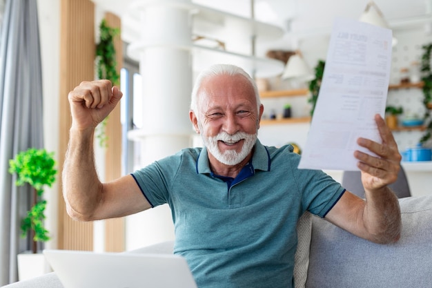 Surpris en riant heureux vieil homme retraité mature regardant à travers un document papier se sentant excité analysant des informations financières obtenant un remboursement d'impôts ou une approbation de prêt bancaire à la maison