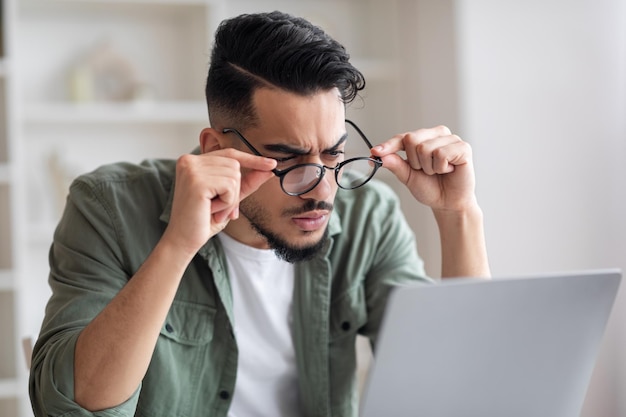 Surpris sérieux jeune homme arabe avec barbe enlève ses lunettes regarde un ordinateur portable dans le bureau à domicile