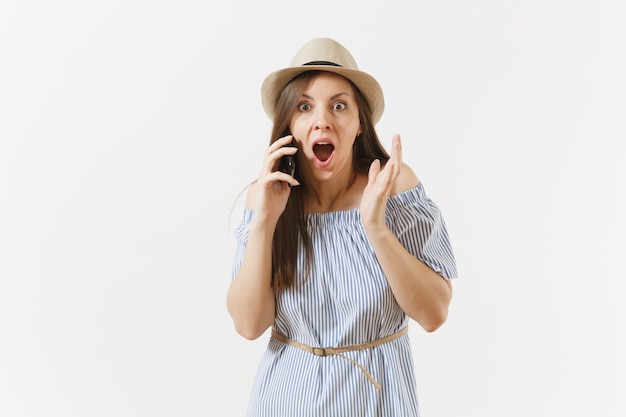 Surprise, bouleversée, jeune femme vêtue d'une robe bleue, le chapeau a des problèmes, entend de fausses nouvelles ou une rumeur inattendue sur un téléphone portable isolé sur fond blanc. Personnes, émotions sincères, concept de style de vie.