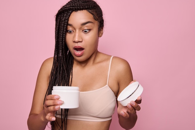 Surprise choquée belle femme africaine ouvrant un pot avec un produit de beauté et le regardant avec étonnement et frustration, isolée sur fond rose avec espace de copie