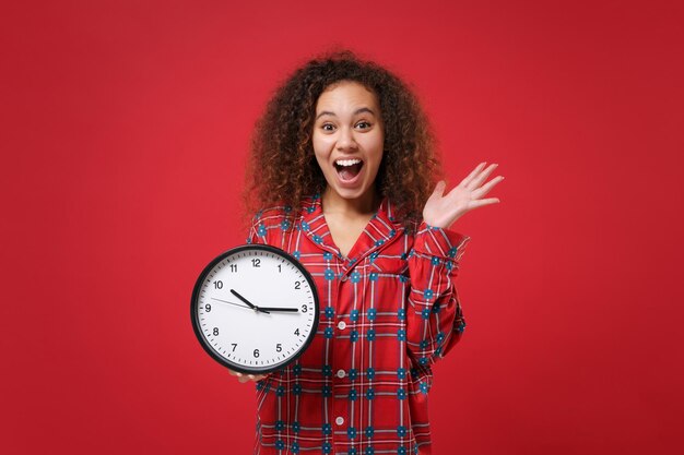 Photo surprise jeune fille afro-américaine en pyjama homewear posant tout en se reposant à la maison isolée sur fond rouge. détendez-vous le concept de mode de vie de bonne humeur. maquette de l'espace de copie. tenez l'horloge en écartant les mains.
