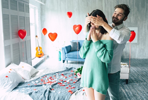 Surprise romantique pour votre bien-aimé. Un bel homme embrasse et ferme les yeux sur sa petite amie ou sa femme en lui faisant un dîner romantique dans la chambre pour leur anniversaire ou la Saint-Valentin