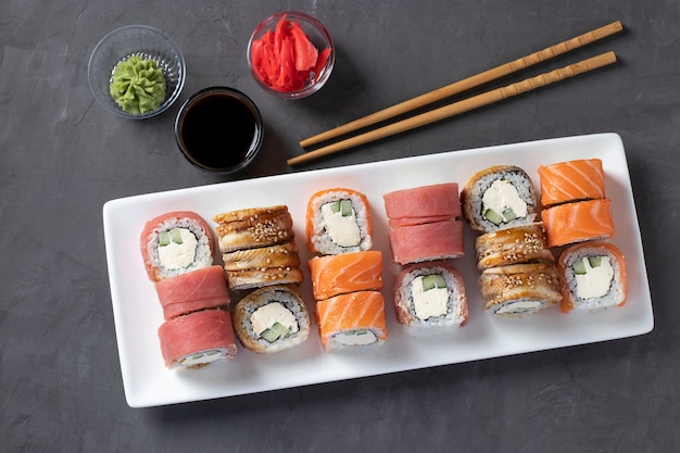 Sushi avec saumon, thon et anguille fumée avec fromage philadelphie sur plaque blanche sur fond gris. Servi avec sauce soja, wasabi, gingembre mariné et bâtonnets pour sushi. Vue d'en-haut