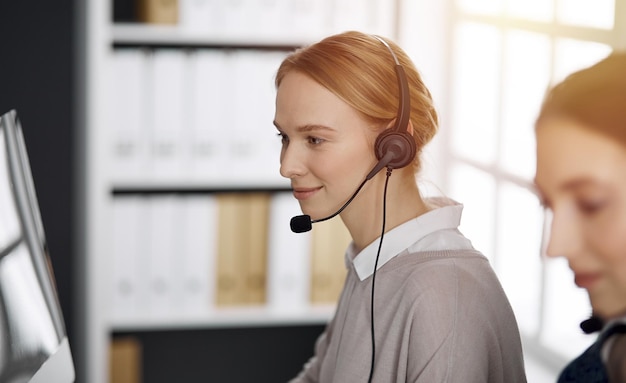 Sympathique femme d'affaires caucasienne parlant par casque dans un bureau ensoleillé. Centre d'appels et groupe de personnes diverses dans les affaires.