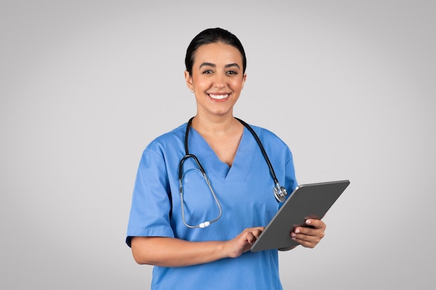 Sympathique jeune femme brésilienne en tenue de travail médicale médecin tenant une tablette numérique et souriant à