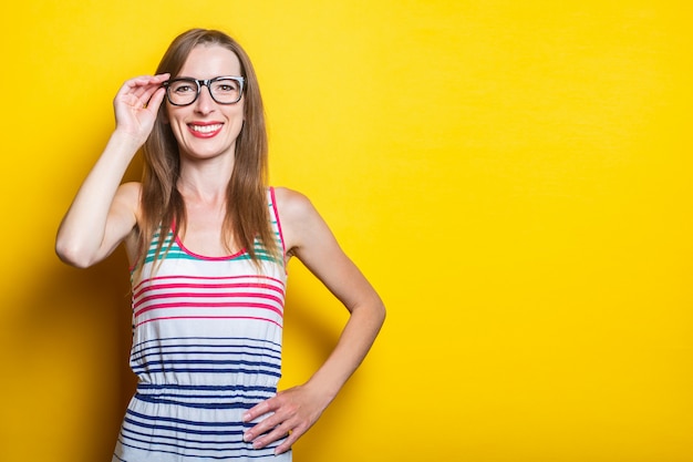 Sympathique jeune fille souriante tenant des lunettes dans une robe rayée sur fond jaune