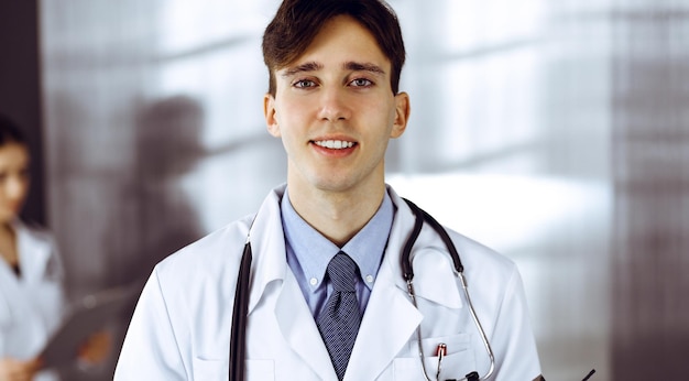 Sympathique jeune homme médecin debout avec presse-papiers dans une clinique moderne. Une collègue est occupée à remplir le formulaire de dossiers de médicaments à l'arrière-plan du médecin. Notion de médecine.