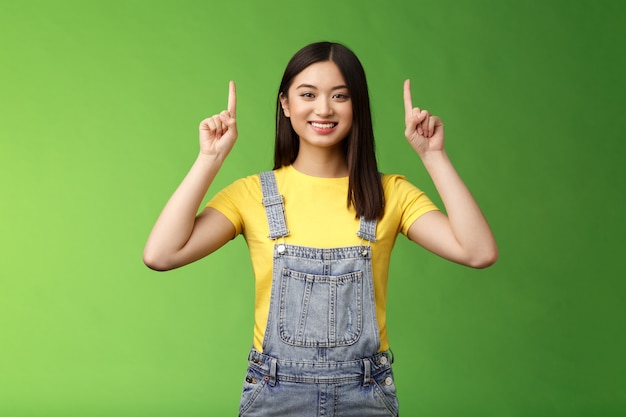 Sympathique jolie femme brune asiatique pointant vers le haut, levez les doigts en haut de la publicité, affiche une promo sûre d'elle, souriante satisfaite, présente un nouveau produit, fond vert.