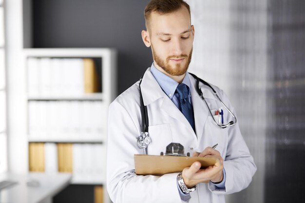 Sympathique médecin à barbe rouge debout et écrivant avec un presse-papiers dans une clinique sur son lieu de travail. Service médical parfait à l'hôpital. Médecine, santé.